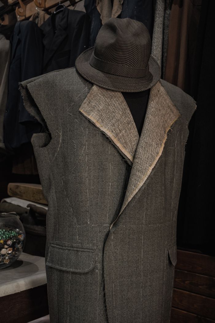 heros-img Close-up of a tailor's mannequin wearing an unfinished suit jacket and hat in workshop.